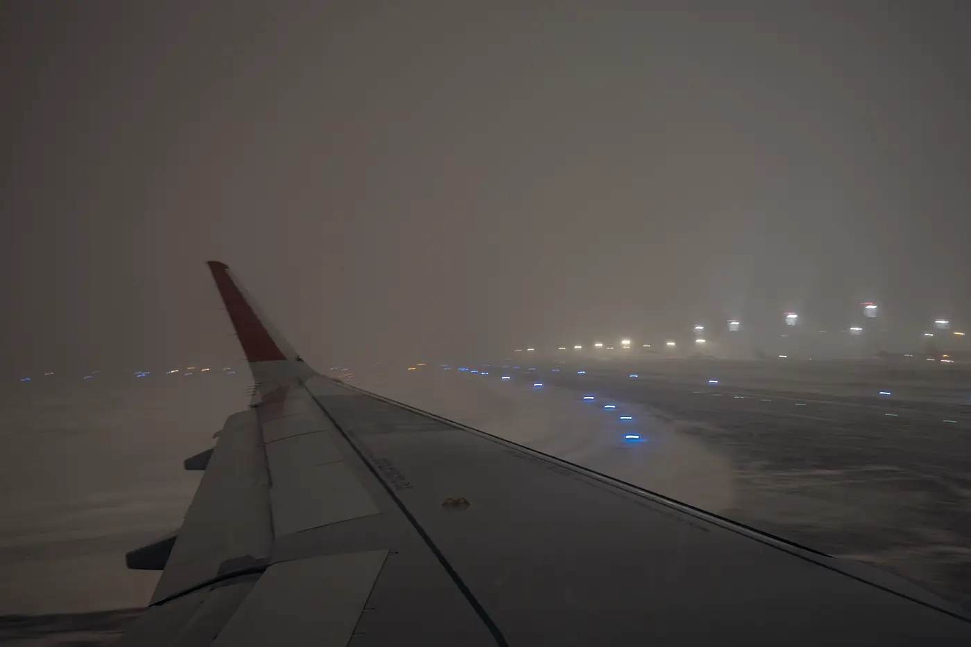 A wing over a snow-covered runway at night, the blue taxiway lights running away into the dark.