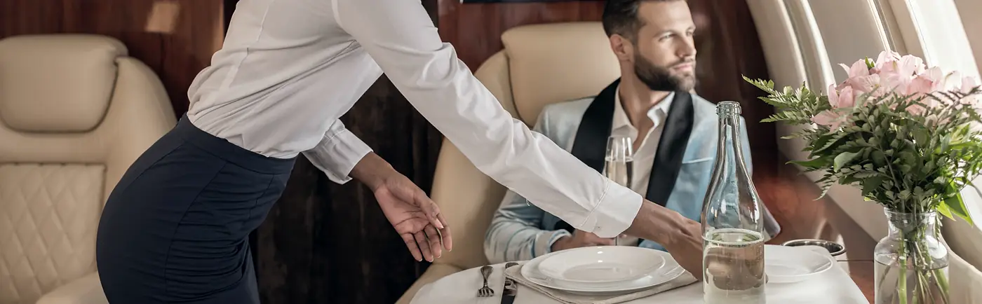 A host laying a table in the cabin, a guest seated beside the window.