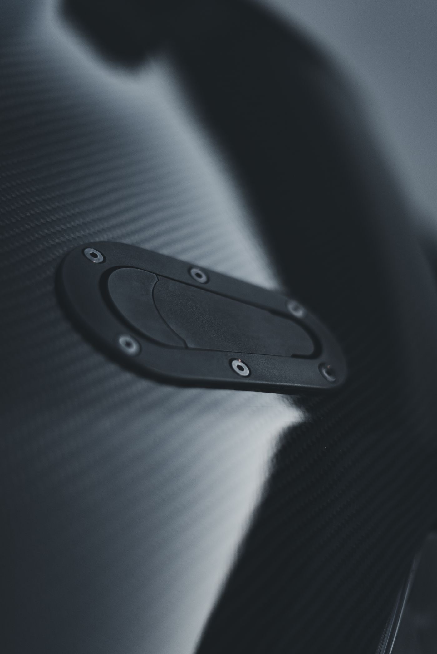 A carbon-fibre panel with a machined access hatch, its weave catching a single studio light.