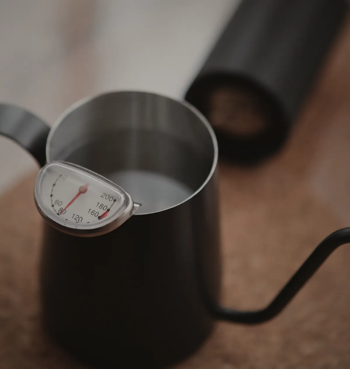 A black gooseneck kettle with a clip-on thermometer, a hand grinder behind it.