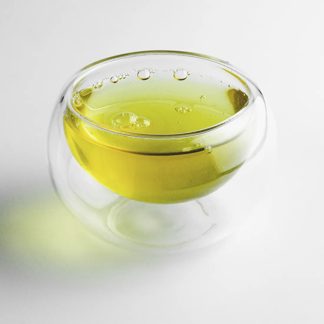 A double-walled glass cup of brewed oolong on a pale grey table: clear yellow-green liquor with light passing through it.