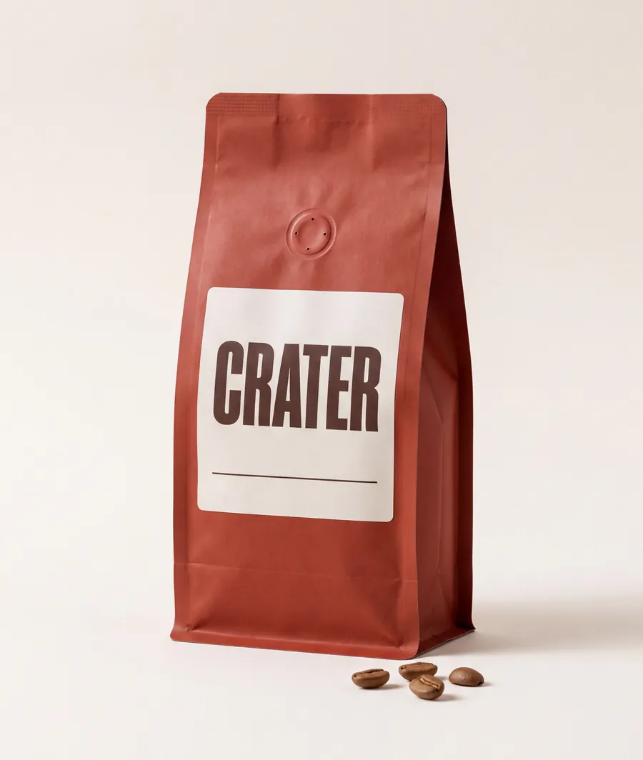 A clay-red paper bag of CRATER coffee with a plain off-white label and a small round valve, standing on a pale backdrop with four roasted beans in front of it.