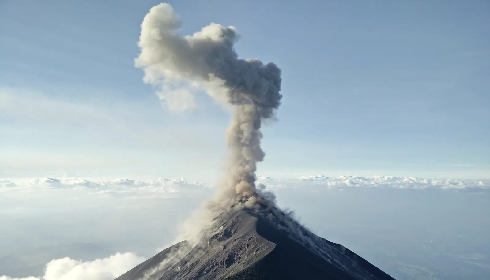 Volcán de Fuego sending a column of grey ash into a clear sky, its cone rising above a layer of cloud.