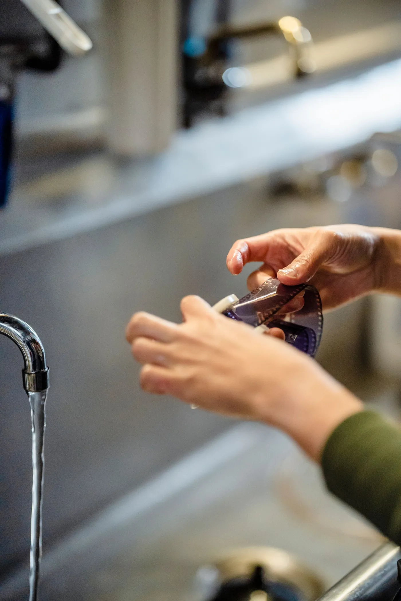 Hands rinsing a strip of film under a running tap at a steel sink.