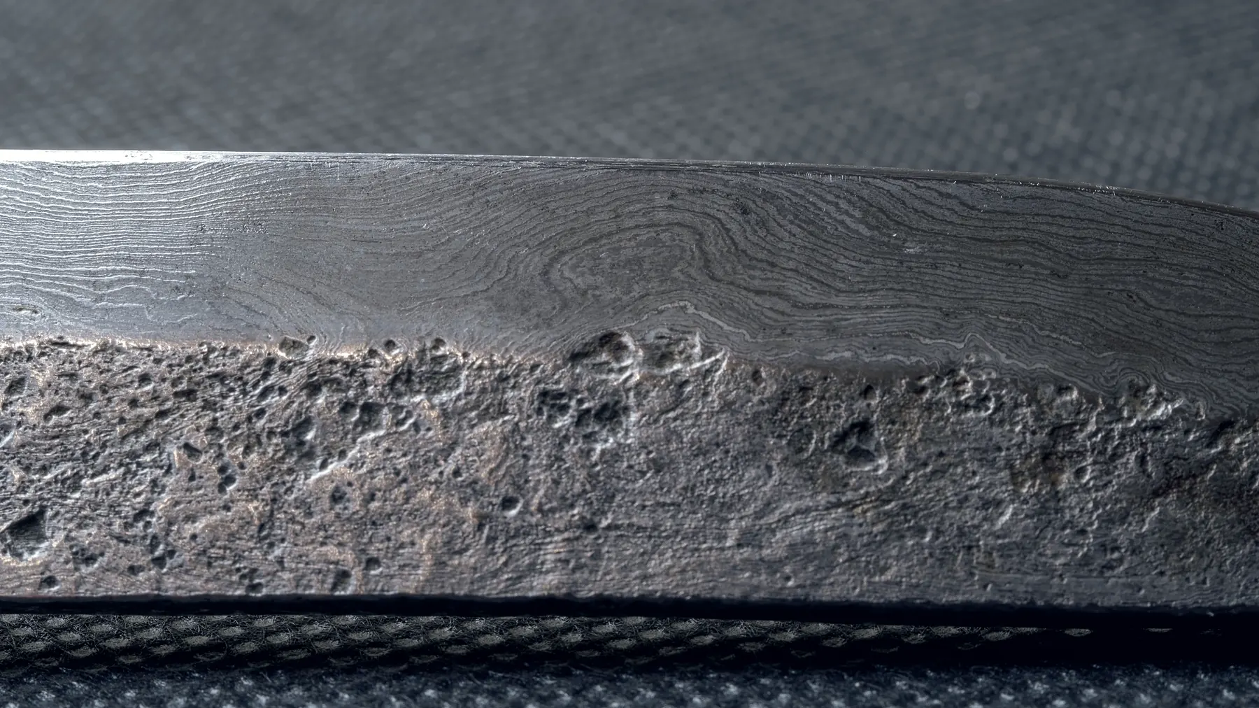 Macro view of a forged blade: fine layered grain running the length of the steel above a rough forge-scale flat.