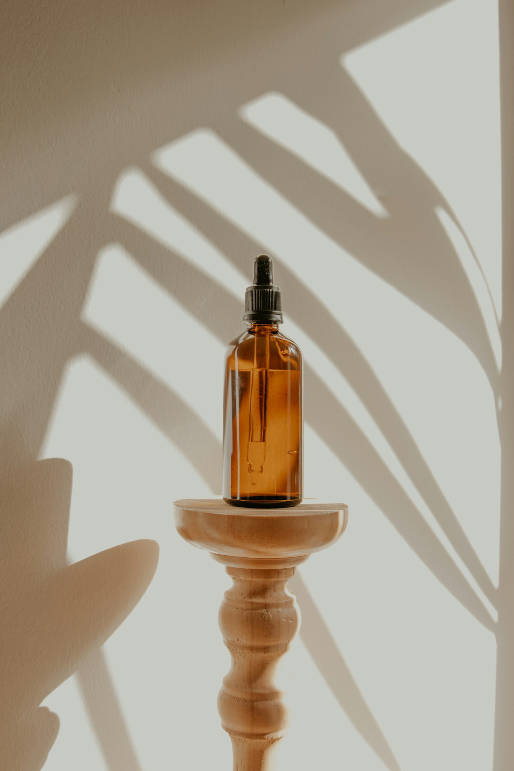 Unlabeled amber dropper bottle on a wooden stand, lit so a leaf shadow falls across the wall.