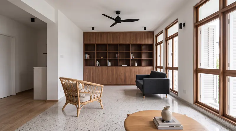 After: a walk-up living room with the original terrazzo floor polished, a walnut bookshelf wall, a rattan chair and white shutters on tall windows.