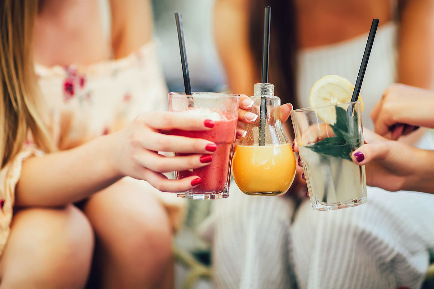 Three hands holding up glasses of pink, orange and pale green drinks with straws