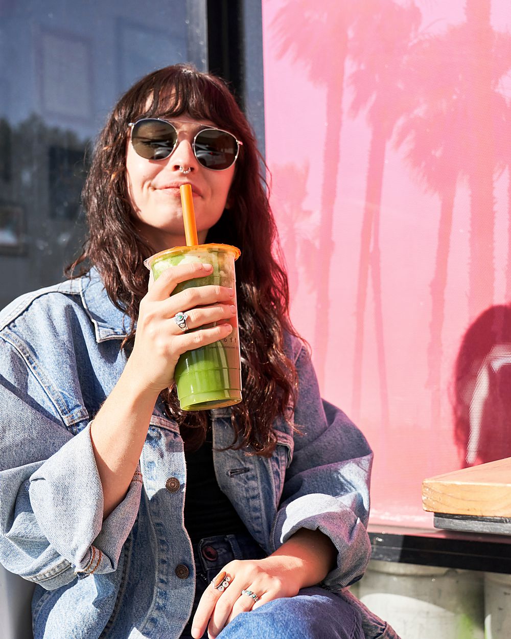A woman in sunglasses and a denim jacket sipping a green drink through a straw in front of a pink wall