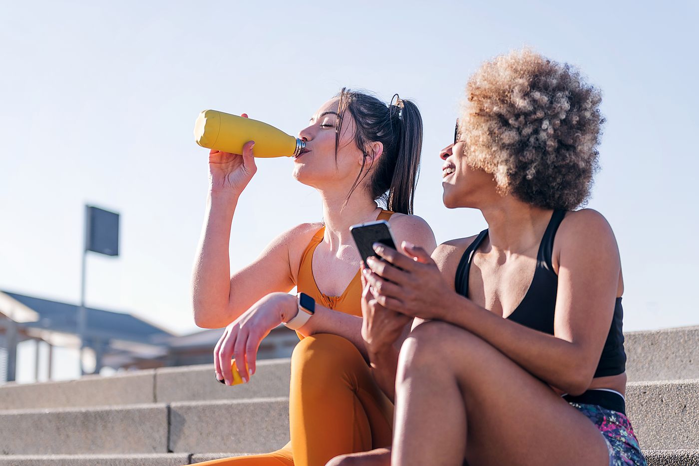 Two women in running clothes sitting on concrete steps, one drinking from a bottle of orange drink