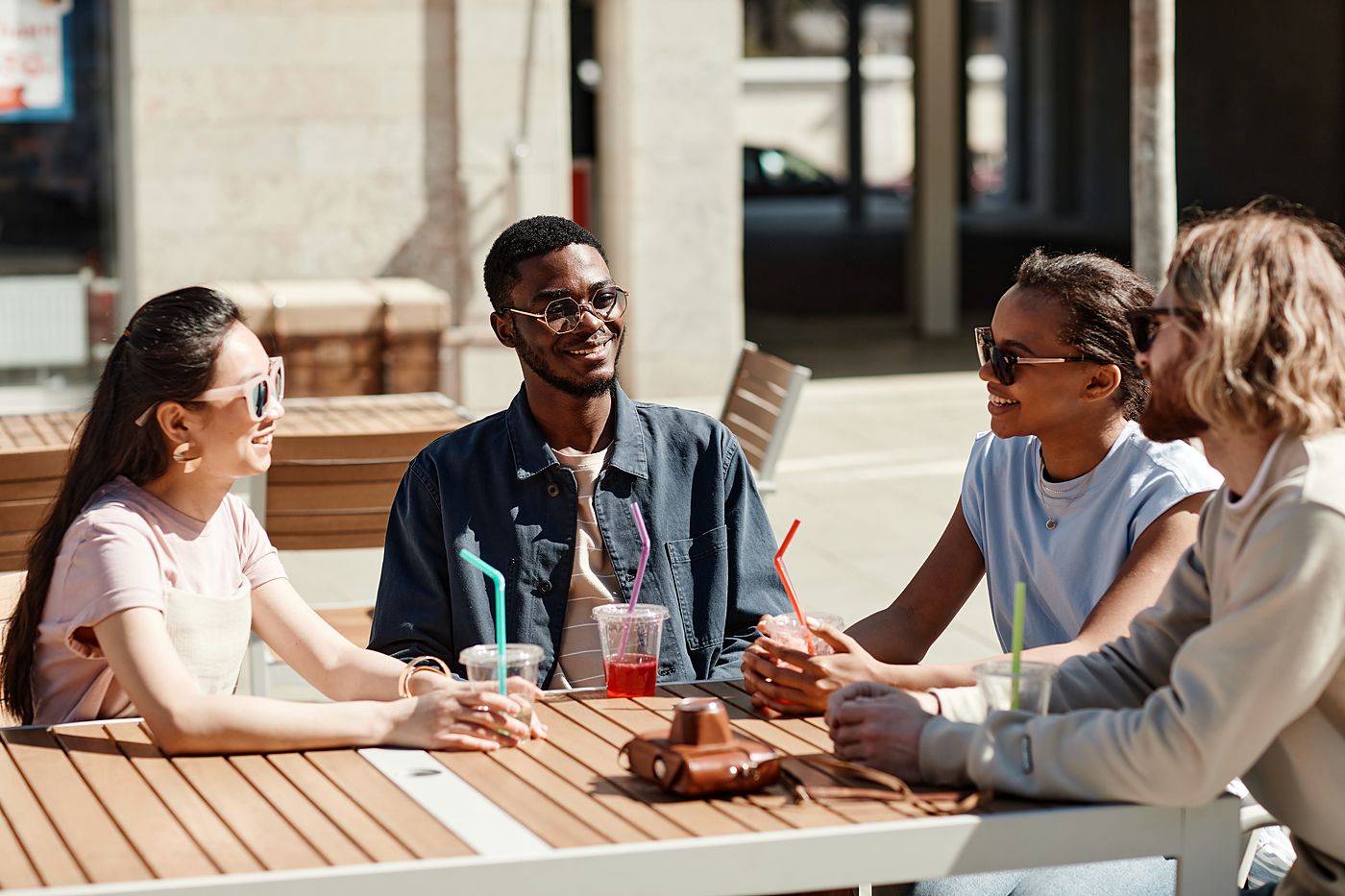 Four friends at an outdoor café table in the sun with coloured drinks in cups and straws