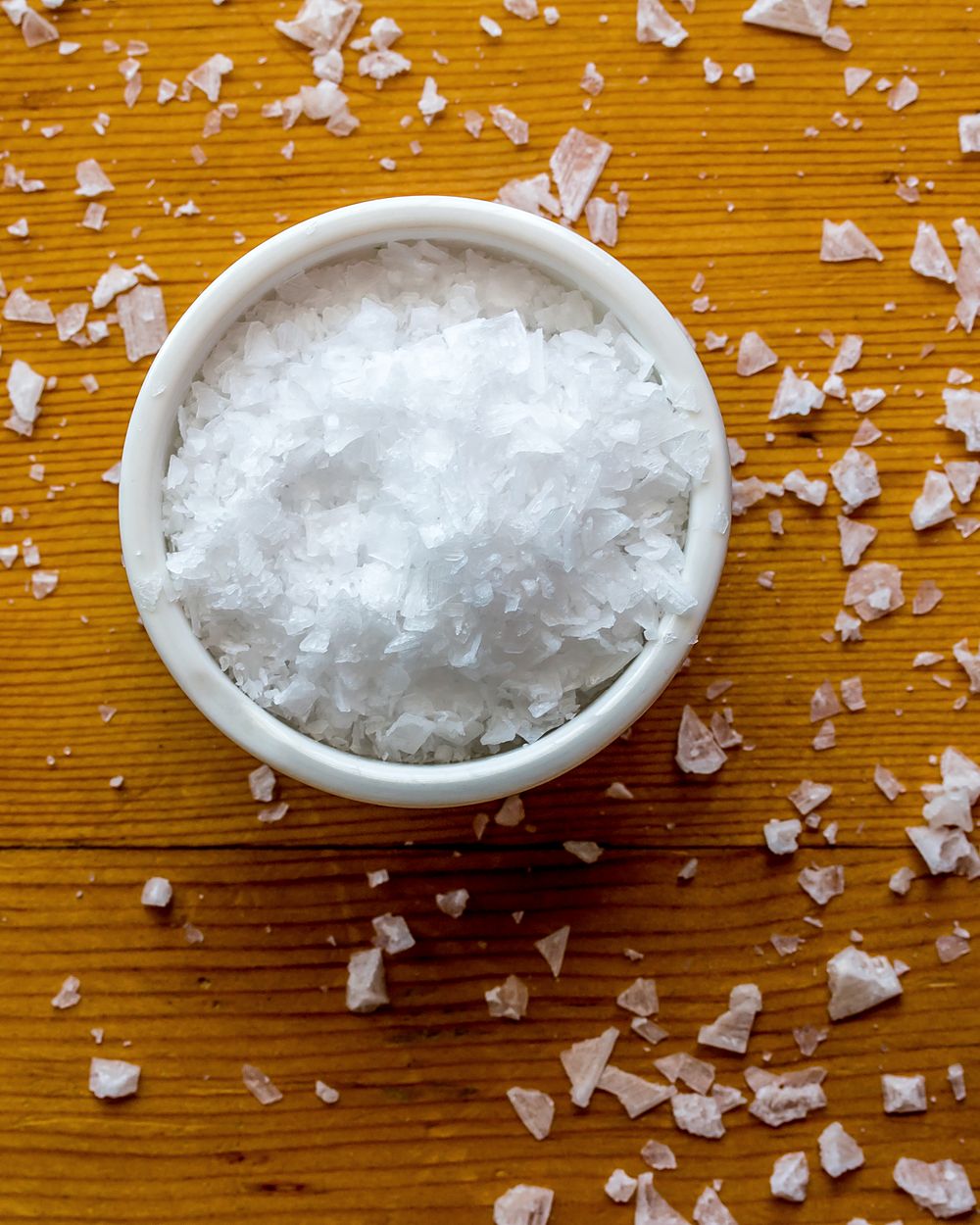 A small bowl of sea salt flakes on a wooden table, a few flakes scattered around it