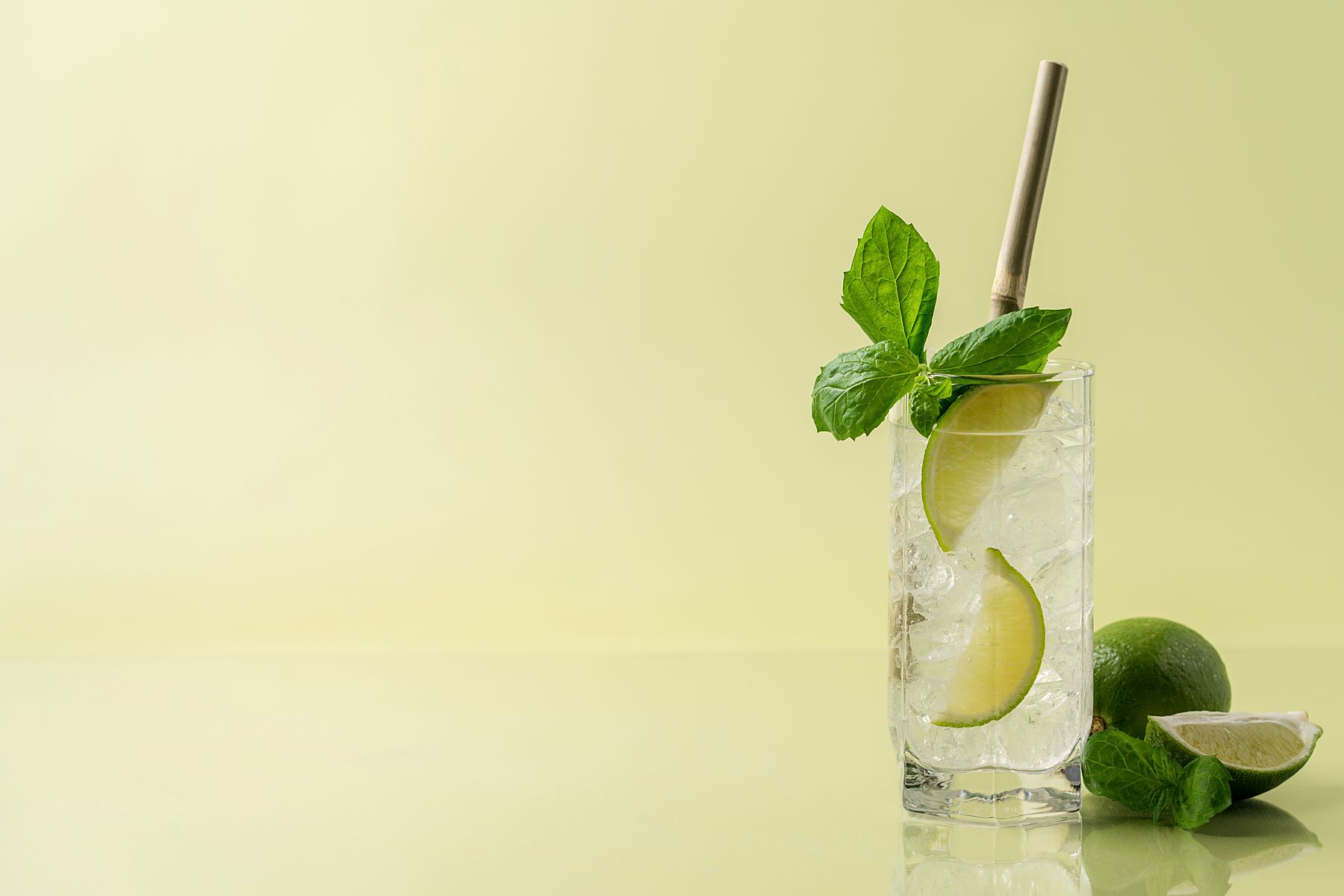A tall glass of pale sparkling drink over ice with a lime wheel and mint, on a pale yellow table