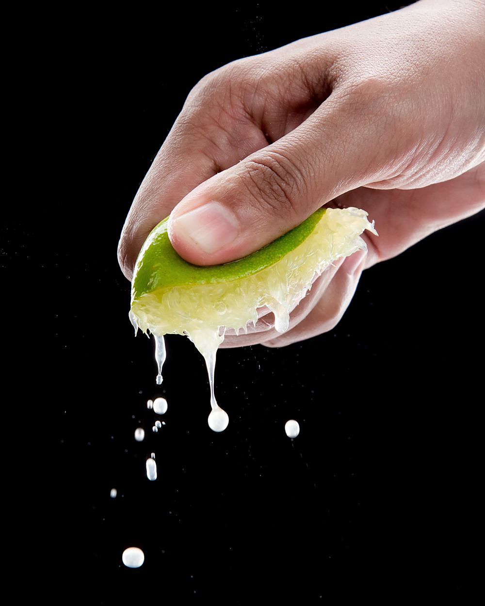 A hand squeezing half a lime, juice falling, against a black background