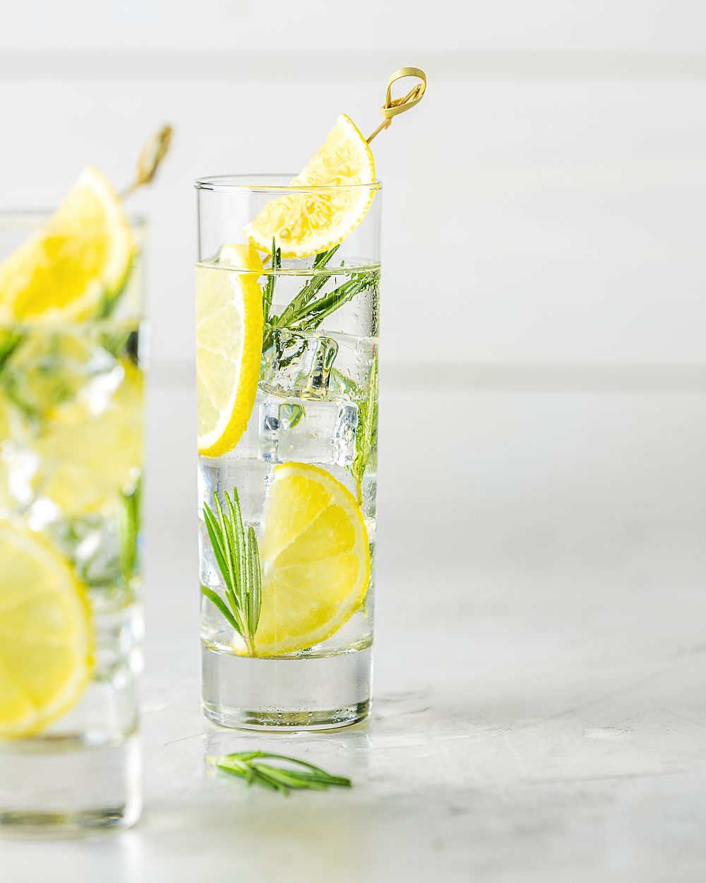 Two tall glasses of sparkling citrus drink with ice and rosemary on a grey table