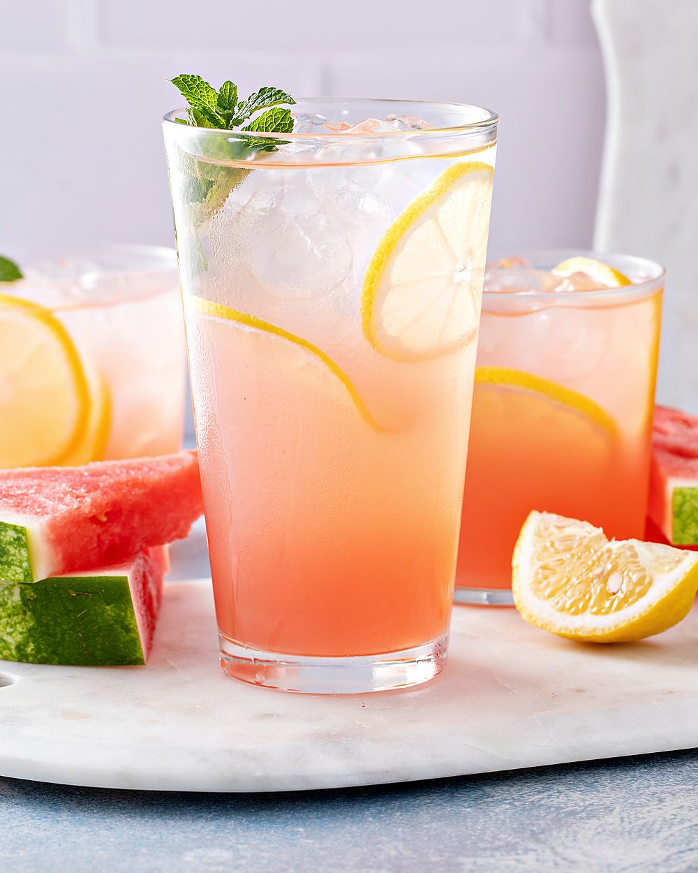 Tall glasses of pink watermelon drink with ice beside wedges of watermelon on a board