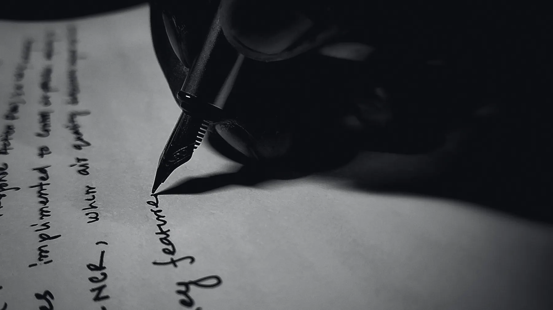 A hand writing a line of cursive with a fountain pen in low light, the nib and the wet ink catching what light there is.