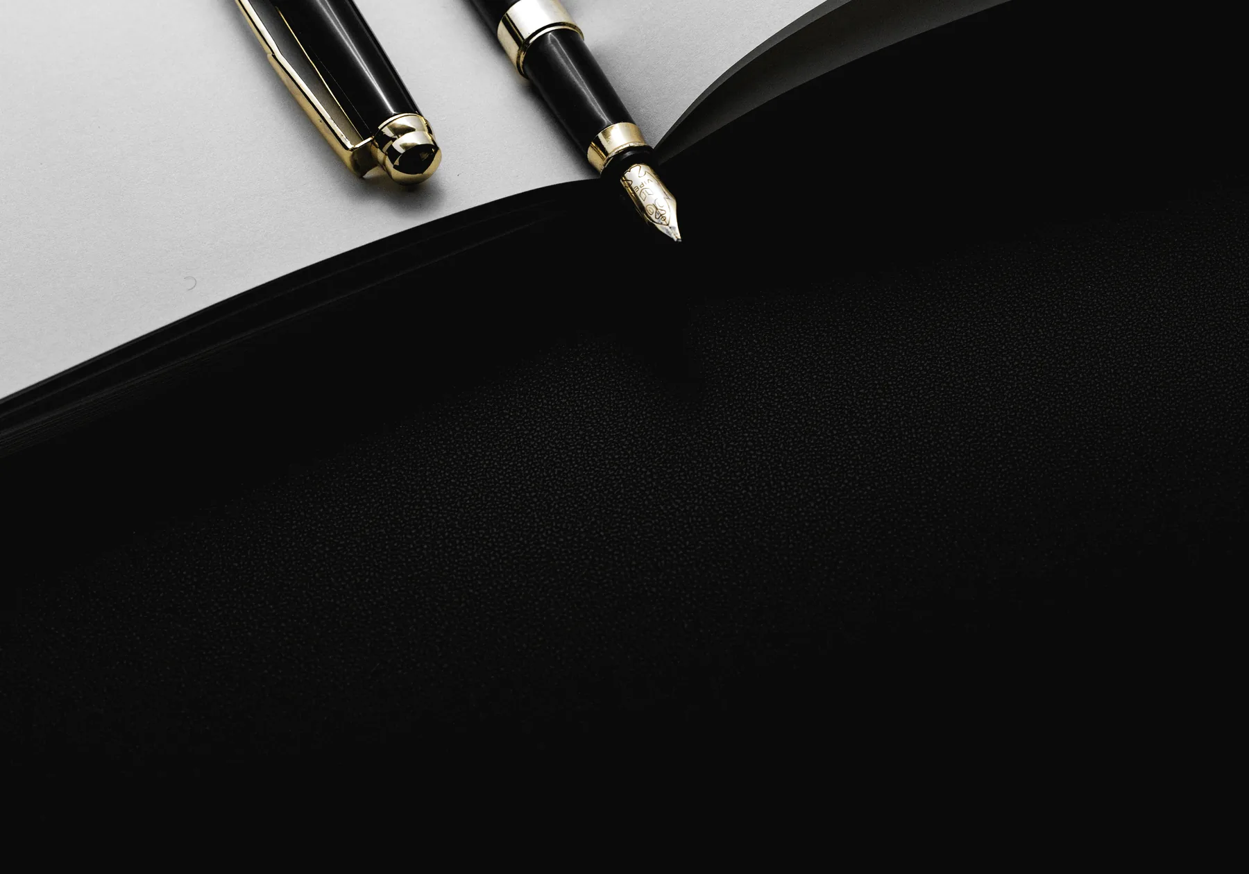 A black pen with gold trim and its cap, lying across the open pages of a notebook on a black leather surface.