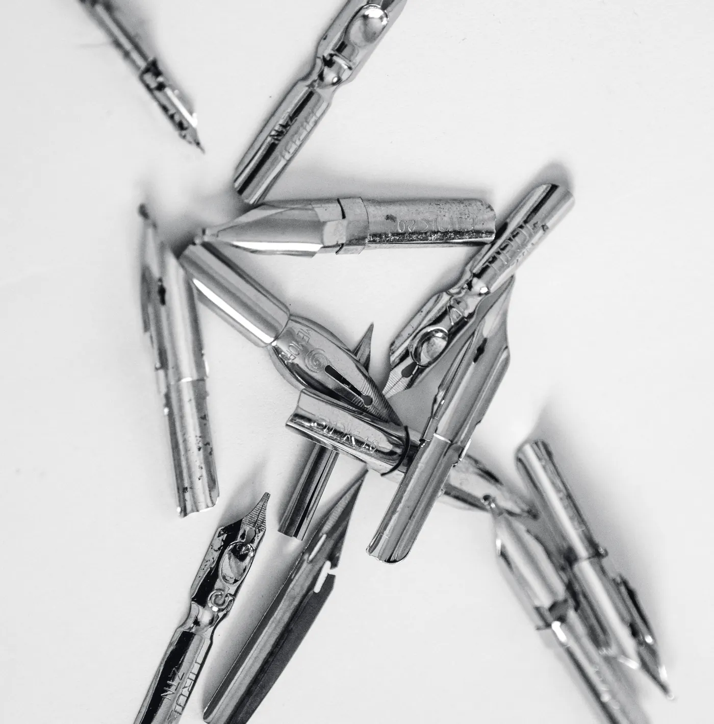 A loose pile of stamped steel dip nibs on a white sheet, in black and white.