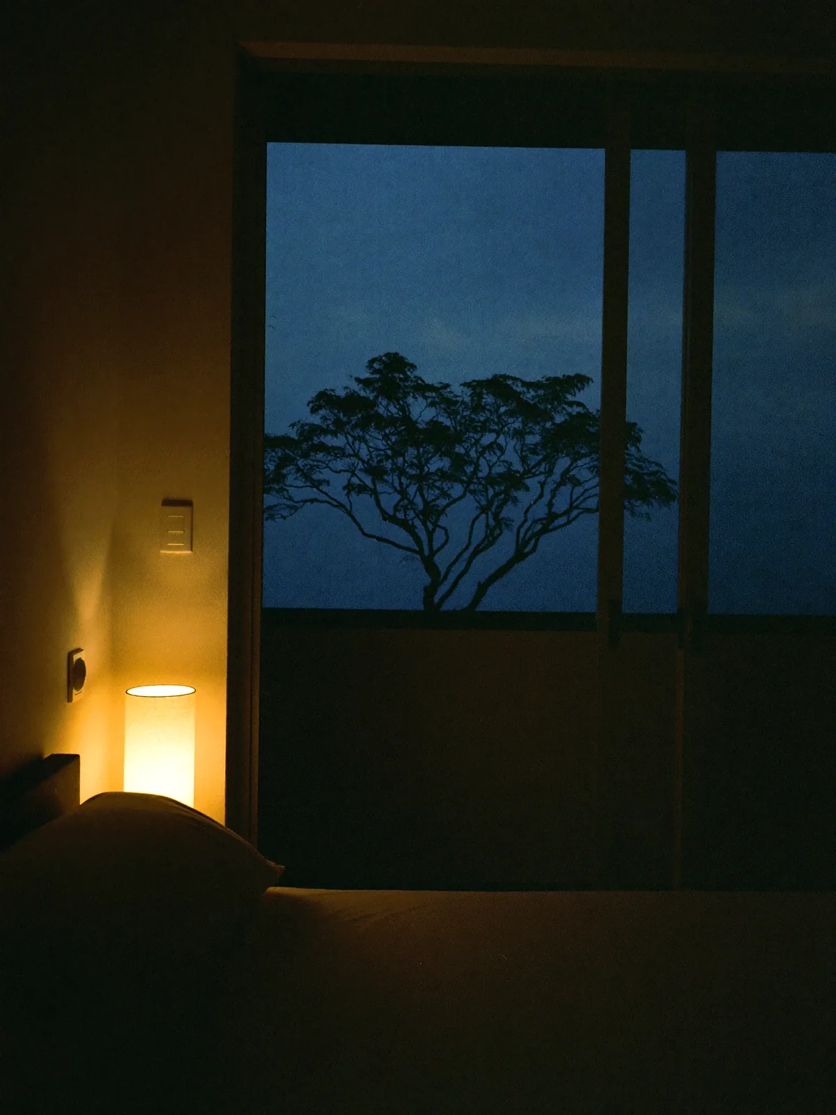 A quiet bedroom at dusk: a low bed under plain linen, one warm bedside lamp, and sliding doors open onto a balcony filled with evening sky and the dark green of a rain tree.
