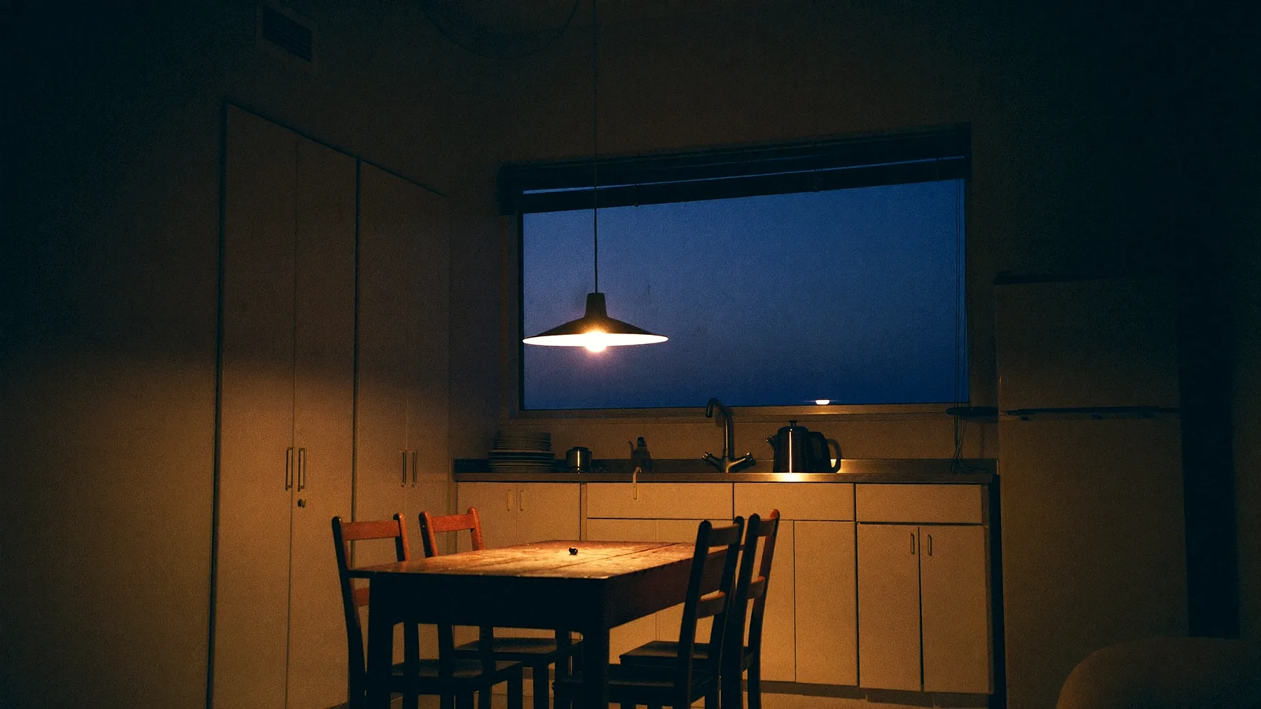 The kitchen and dining end of a flat at dusk: white cabinetry, a worn timber table with mismatched chairs under one warm pendant lamp, and a window of deep blue evening above the sink.