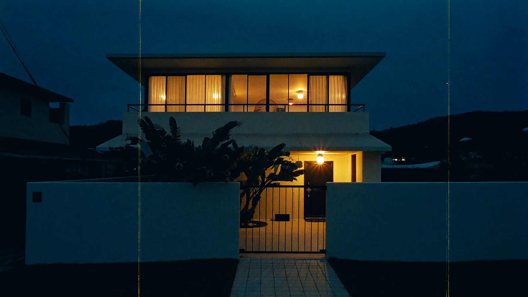 The frontage of a two-storey terrace house at dusk: a low wall and gate, a frangipani in the yard, and a warm porch light beside the door.