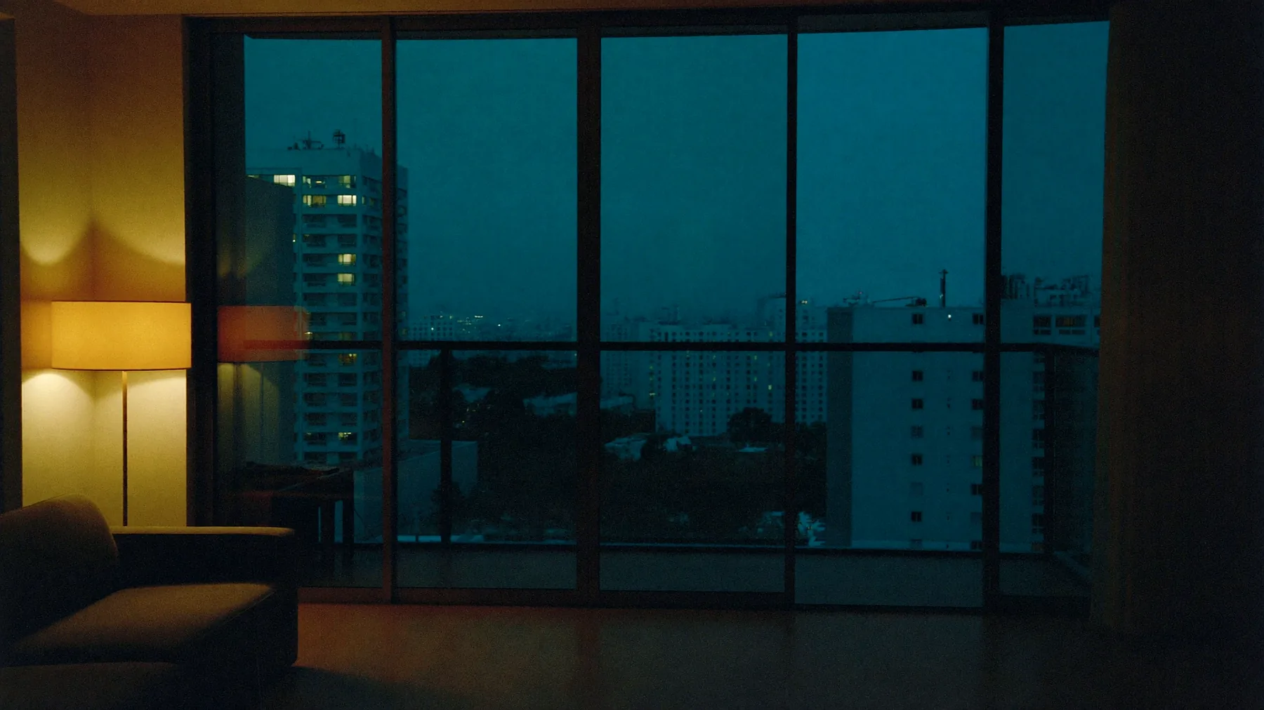 A high-floor living room at dusk: pale oak floor, a low linen sofa, one standing lamp, and a glazed wall onto a balcony above distant lit towers.