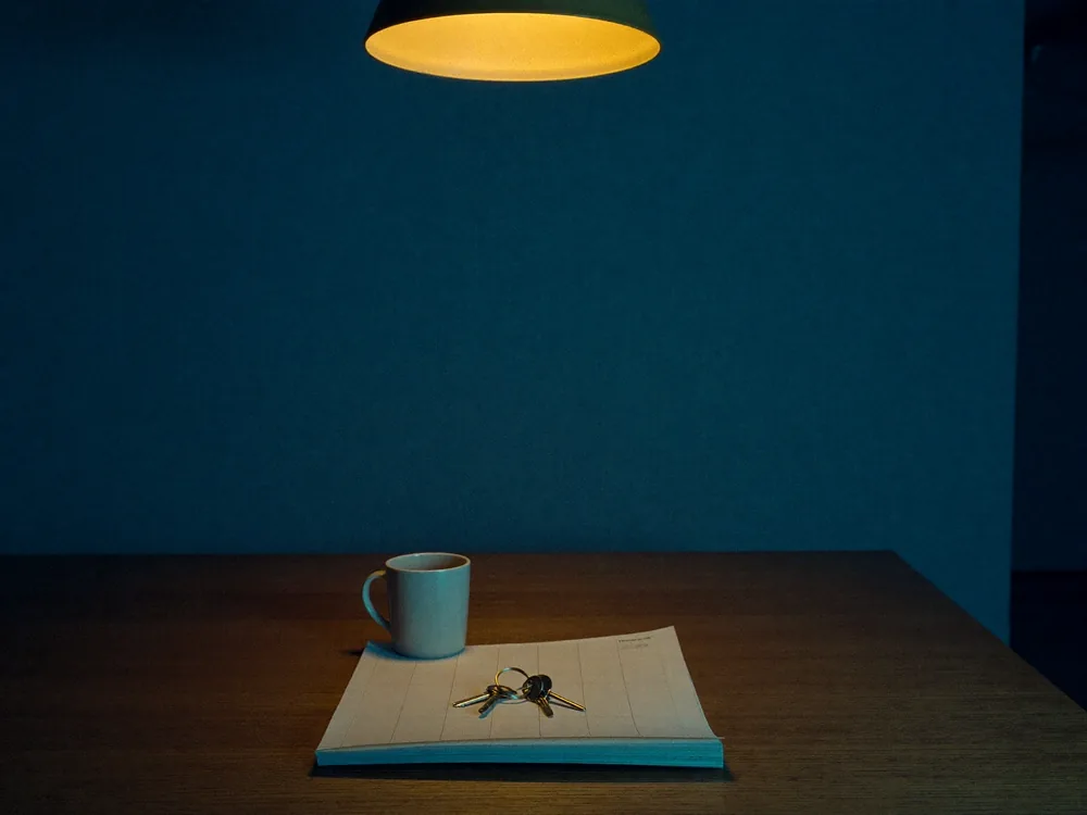 A ring of house keys resting on a folded plan beside a plain white mug, on a bare wooden table under a pendant lamp.