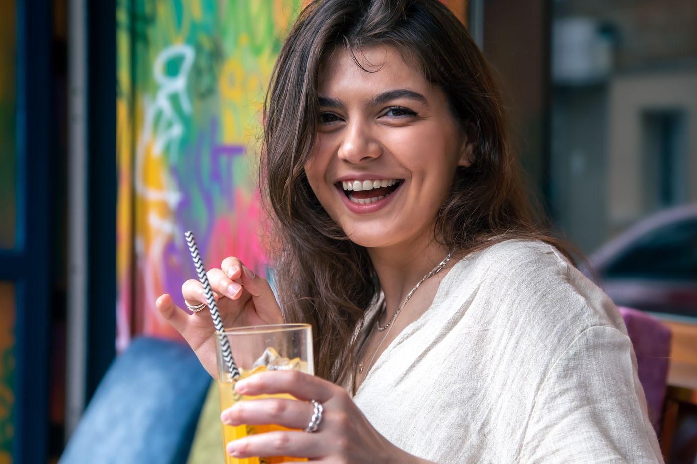 A laughing woman holding a glass of amber drink with a straw, in front of a painted wall.