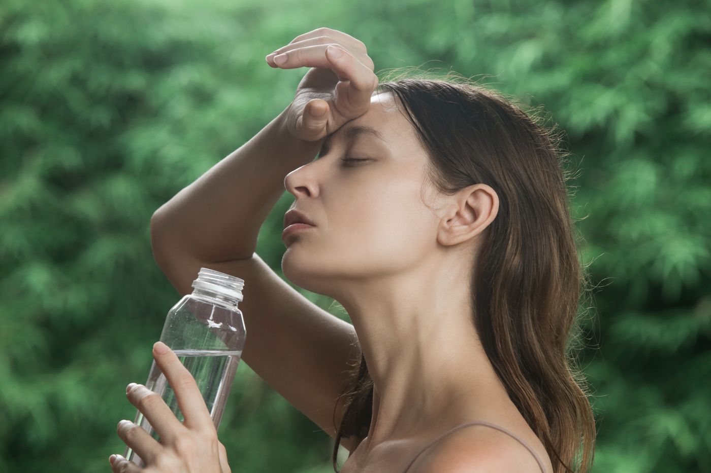 A woman outdoors in the heat, eyes closed, one hand on her forehead, a bottle in the other.