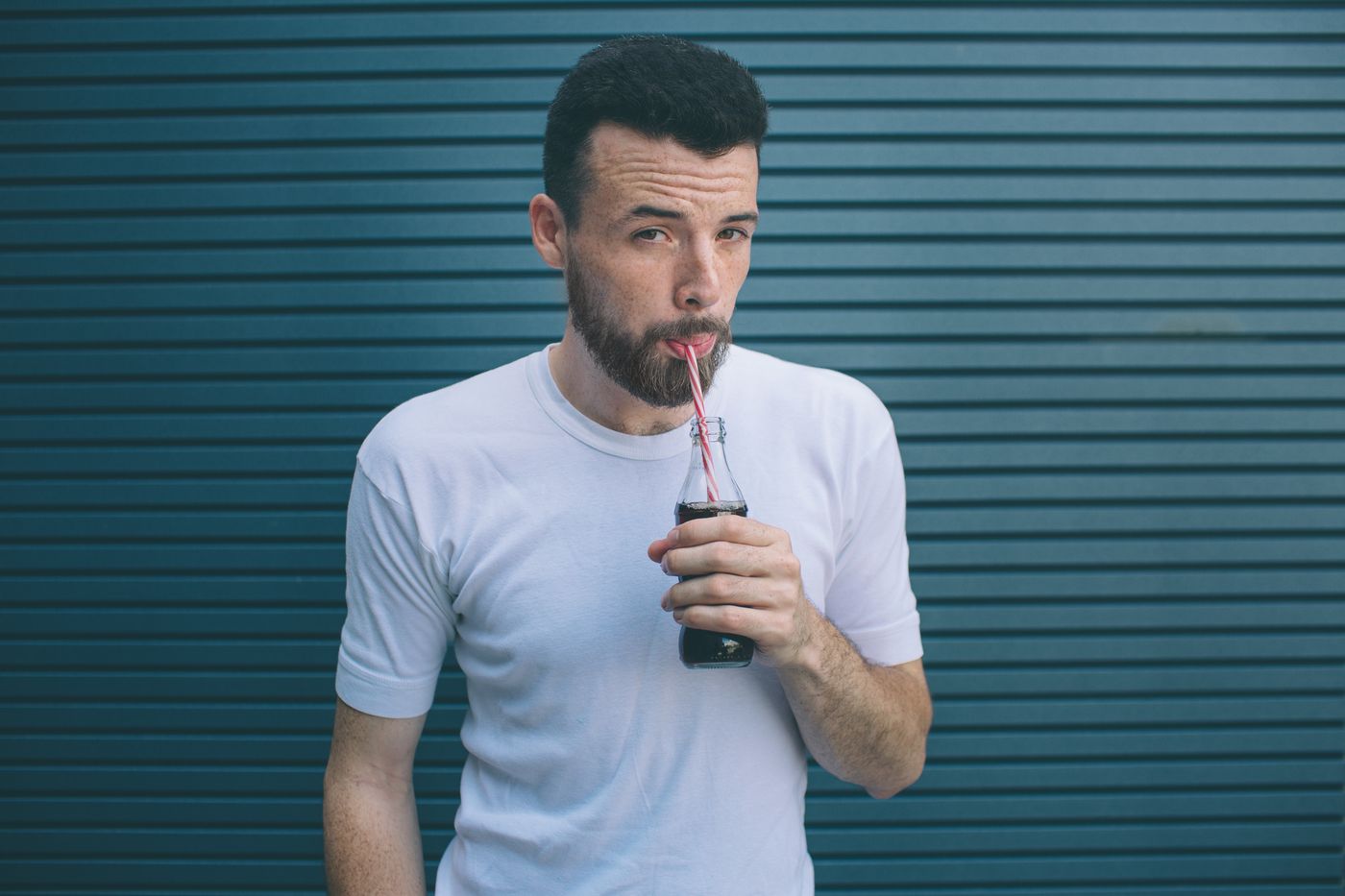 A man in a white t-shirt against a blue corrugated wall, drinking from a glass bottle through a straw, one eyebrow raised.