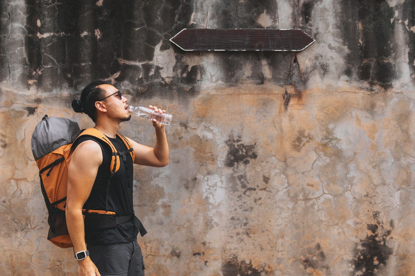 A man with a backpack drinking from a bottle in front of a weathered concrete wall on a hot street.