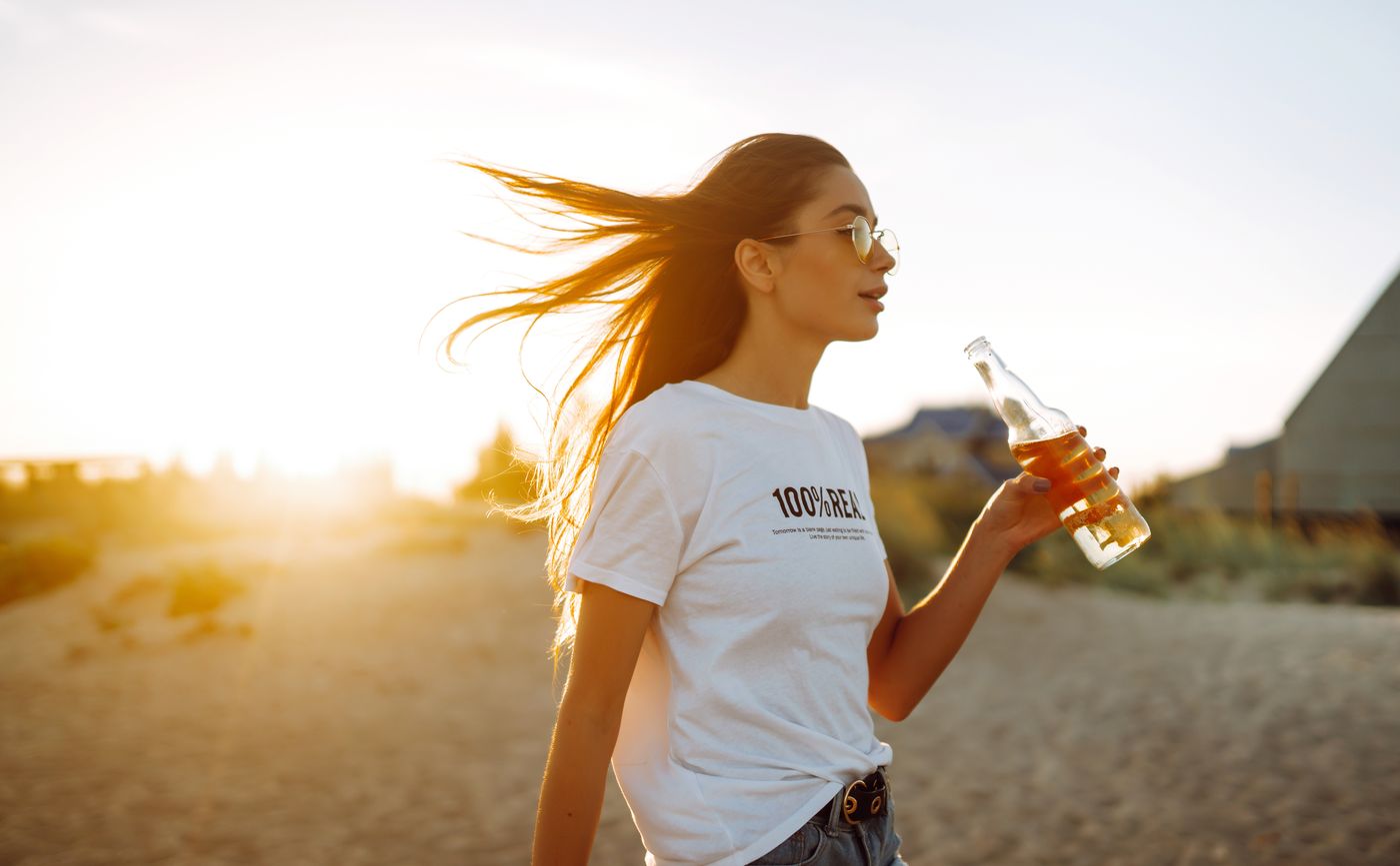 A young woman on a beach at sunset, backlit, drinking from a bottle with her hair blowing across her face.
