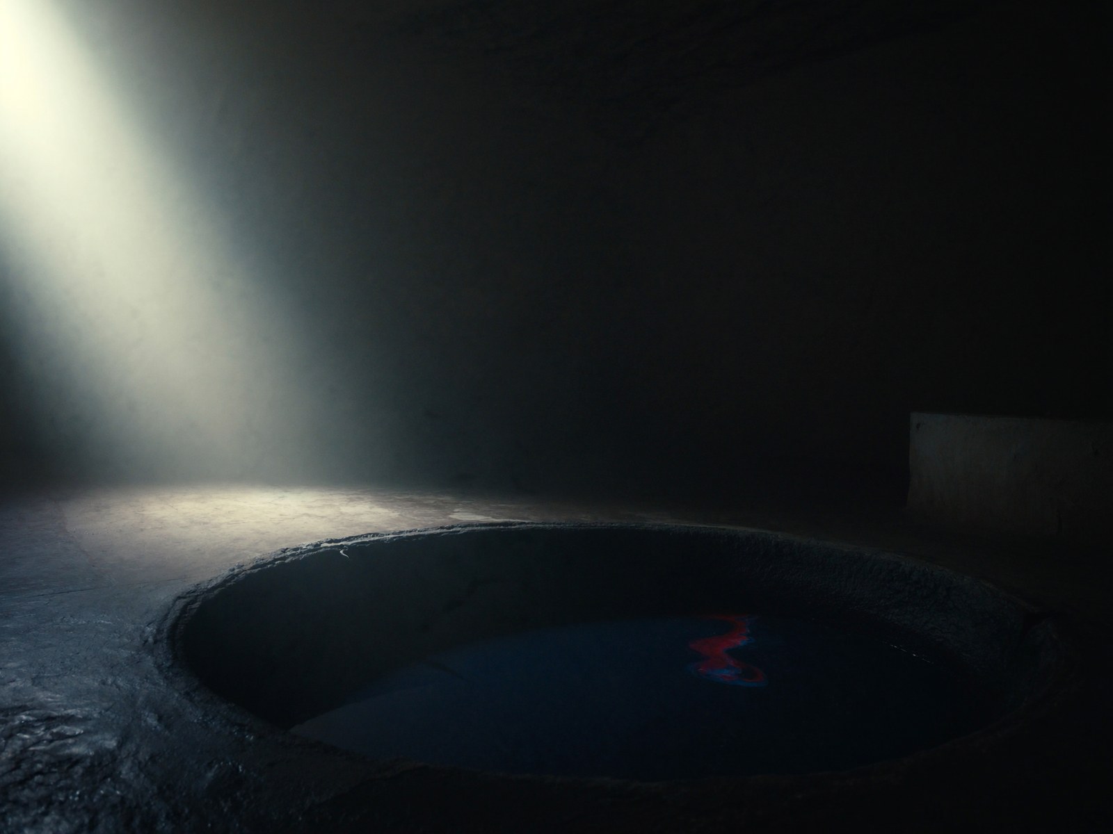 A dye vat sunk into a dark stone floor, one shaft of pale light falling across still indigo water