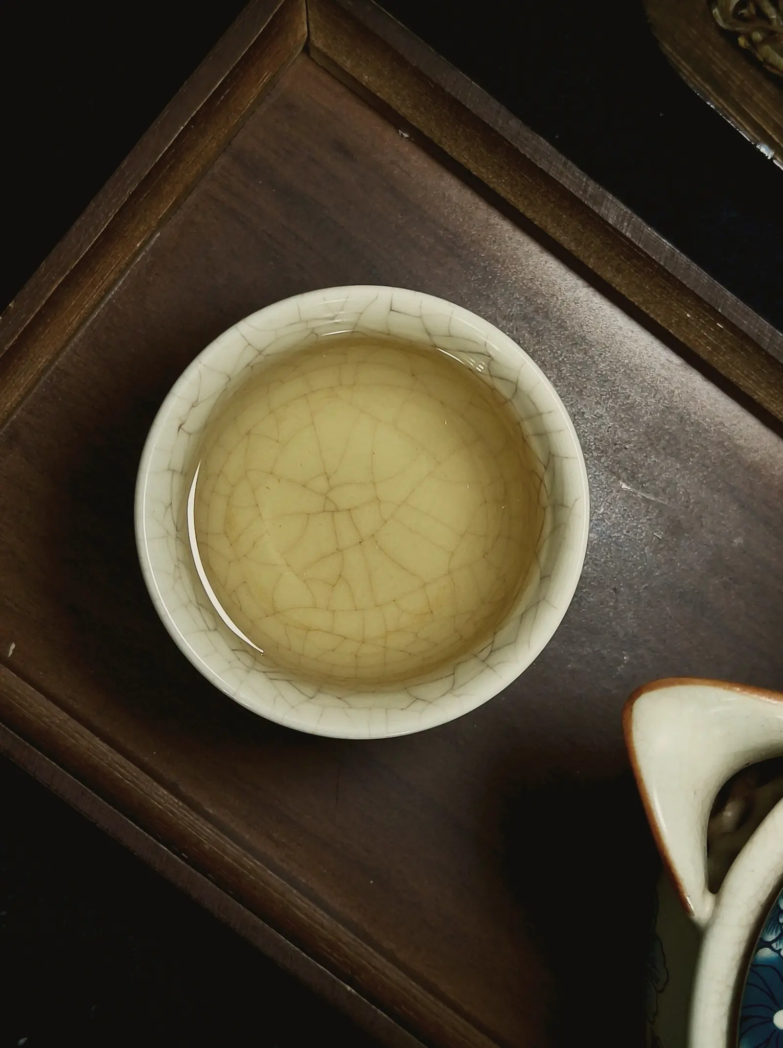 A white cup of pale gold oolong from above, its glaze crackled, on a dark wooden tray