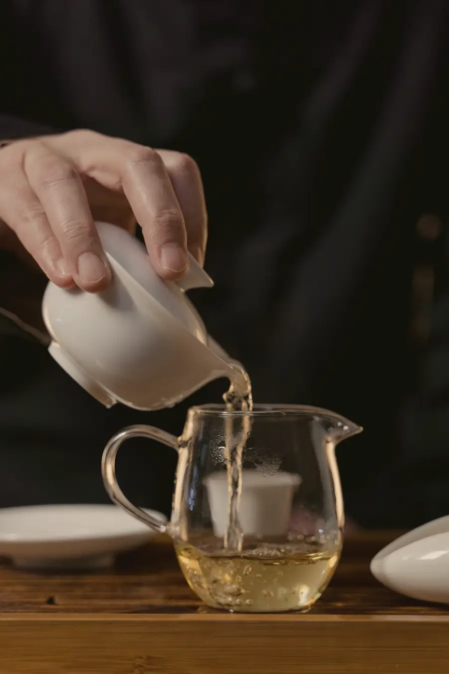A white gaiwan tipped over a glass pitcher, pale gold tea running through a small strainer
