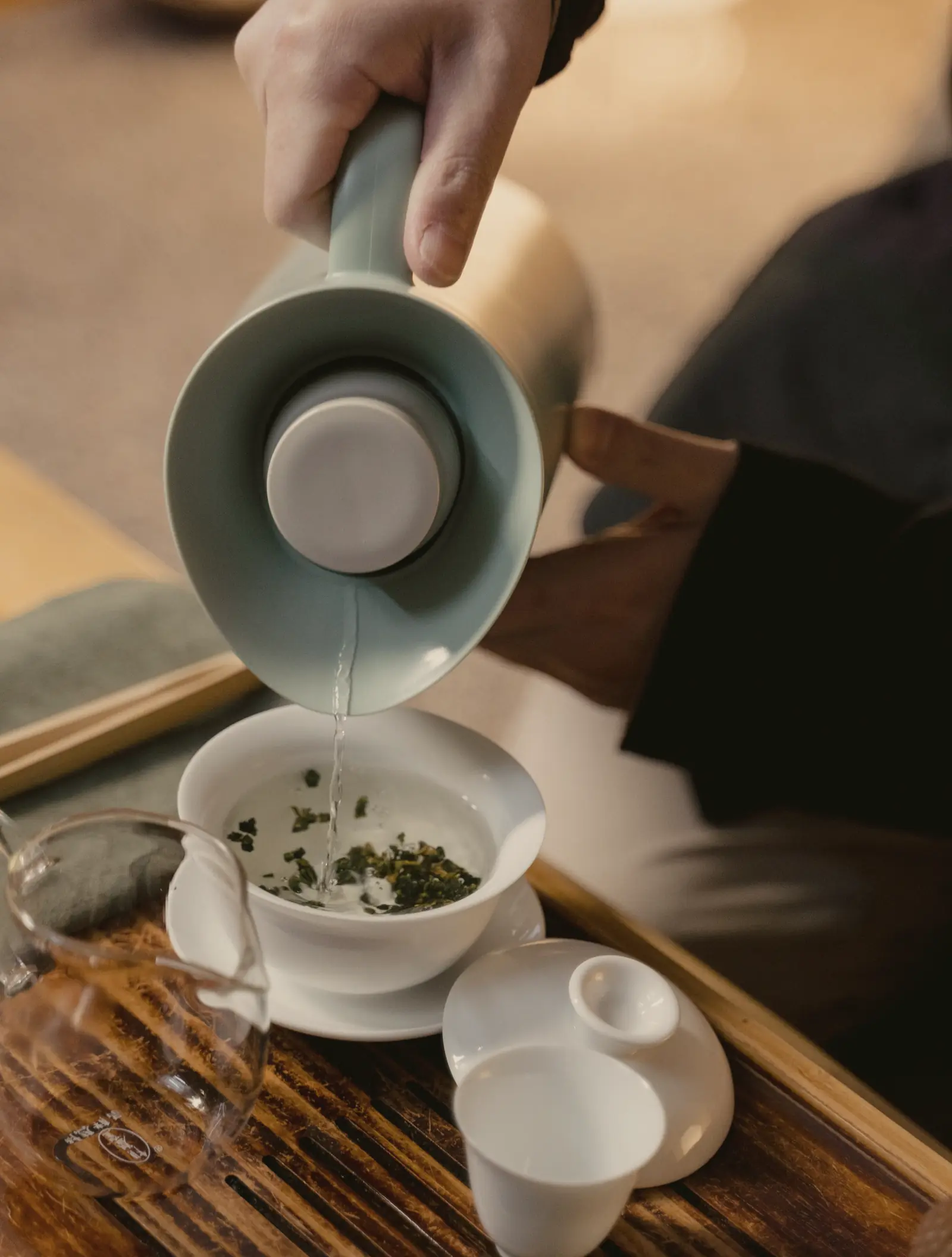 A hand pours water from a pale celadon kettle into a white gaiwan holding rolled oolong, on a slatted tea tray