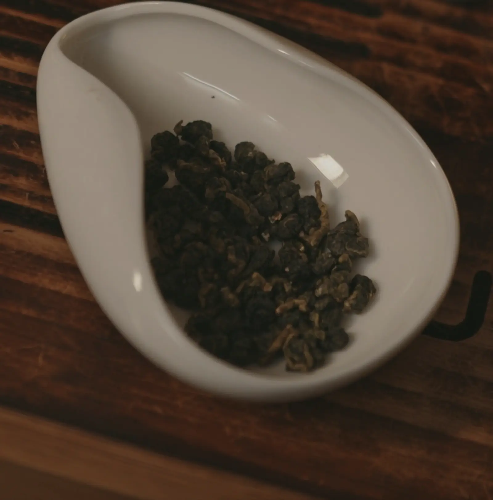 A white porcelain tea scoop holding a heap of dark green rolled oolong knots, on a slatted wooden tea tray
