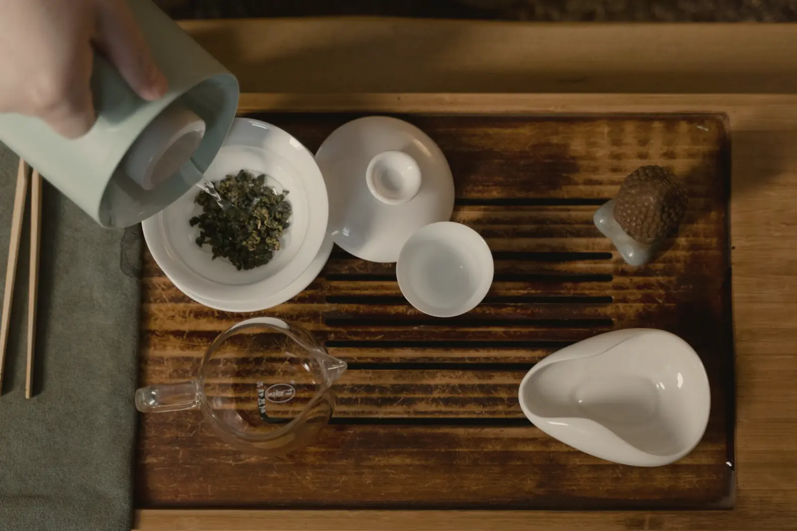 From above: a hand pours from a celadon kettle into a white gaiwan of rolled oolong on a slatted wooden tray, with cups and a glass pitcher