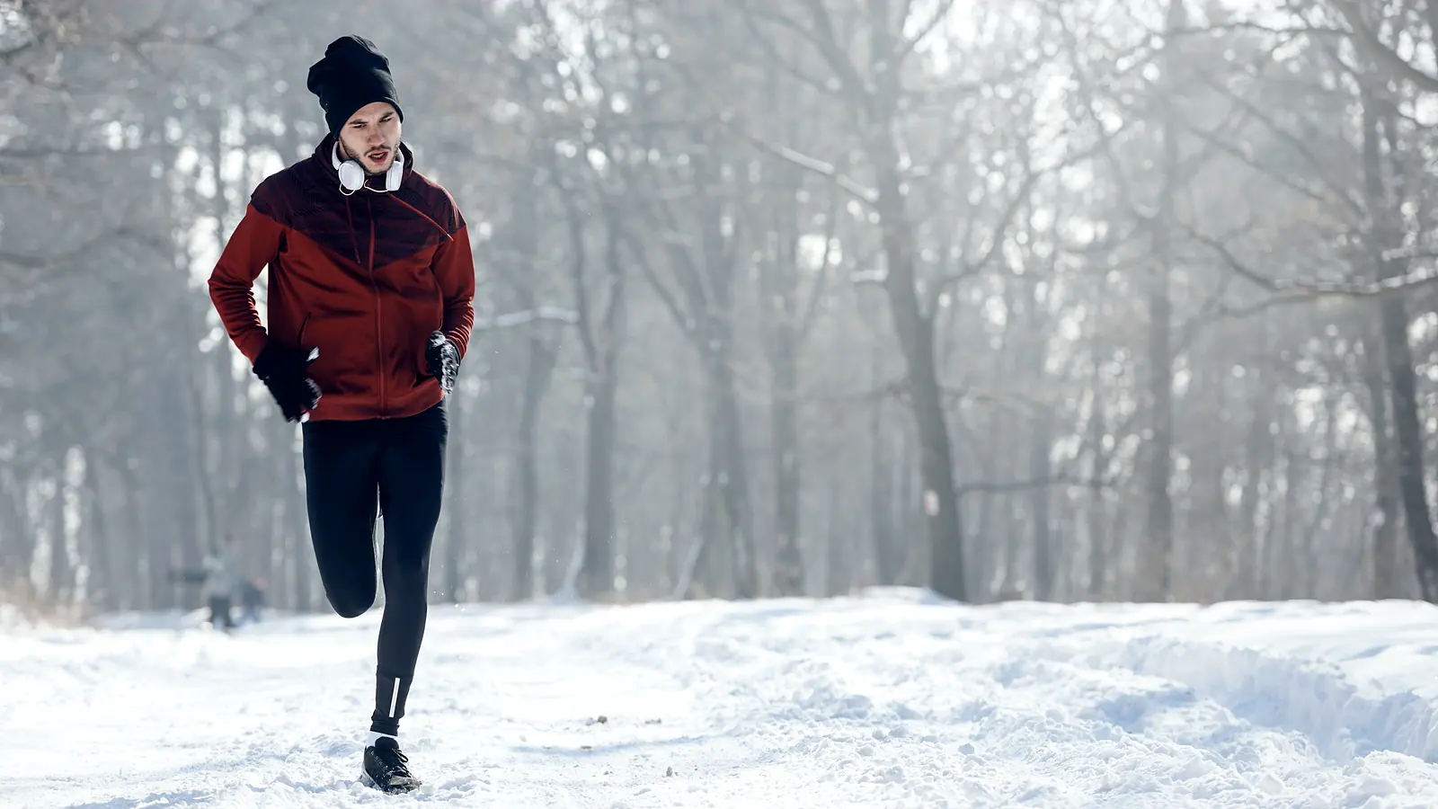 A runner in a shell jacket and tights crossing deep snow below a pine forest.