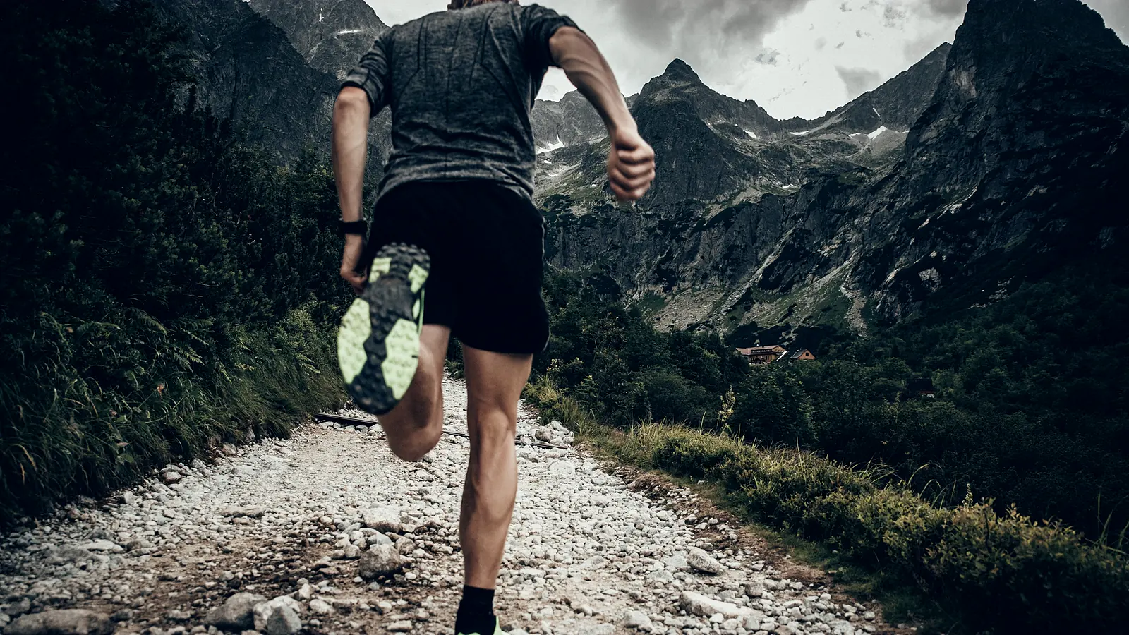 A runner seen from behind on a gravel track climbing towards a grey mountain ridge.
