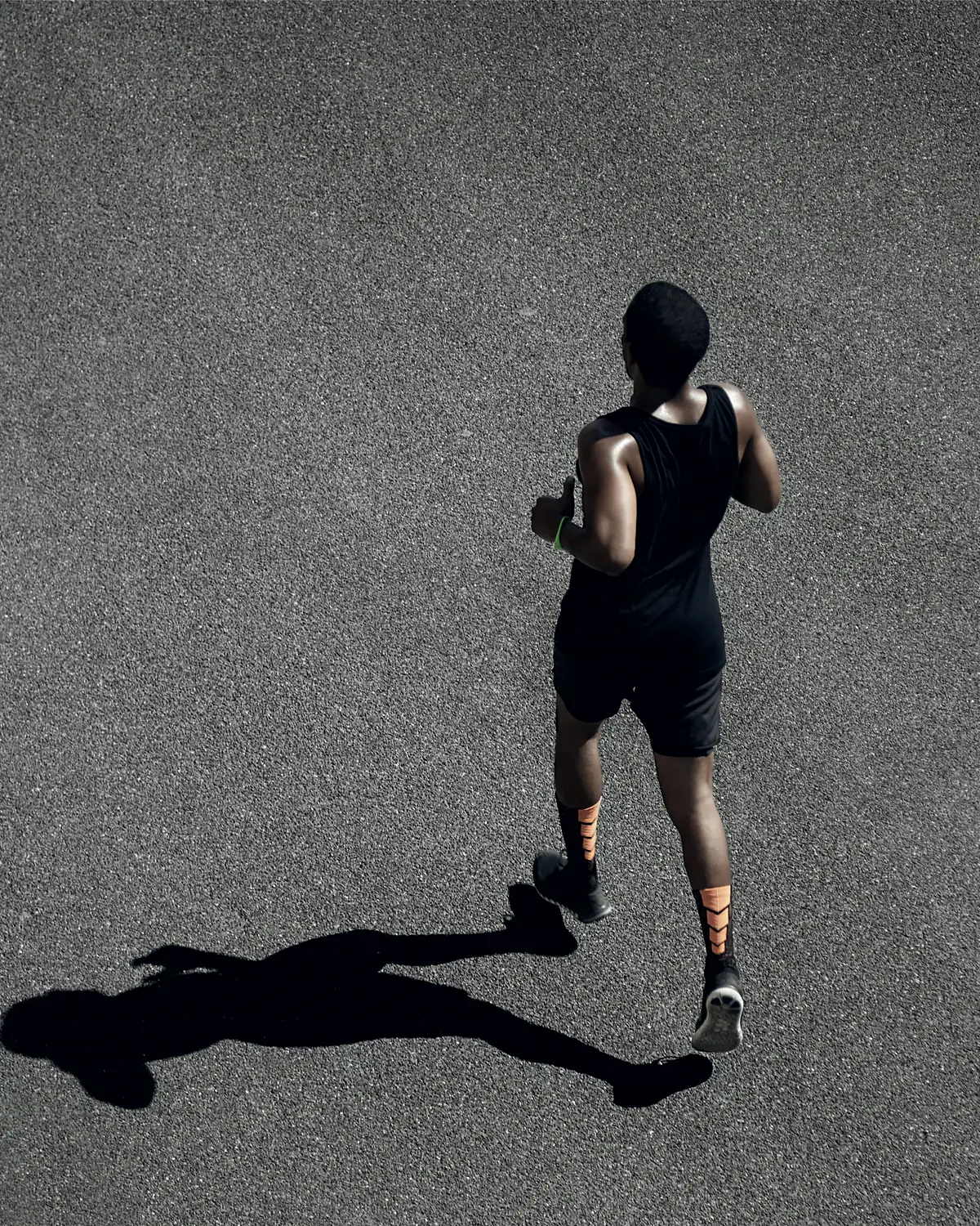 A runner photographed from directly overhead, in black kit on grey asphalt, with a long shadow thrown across the road.