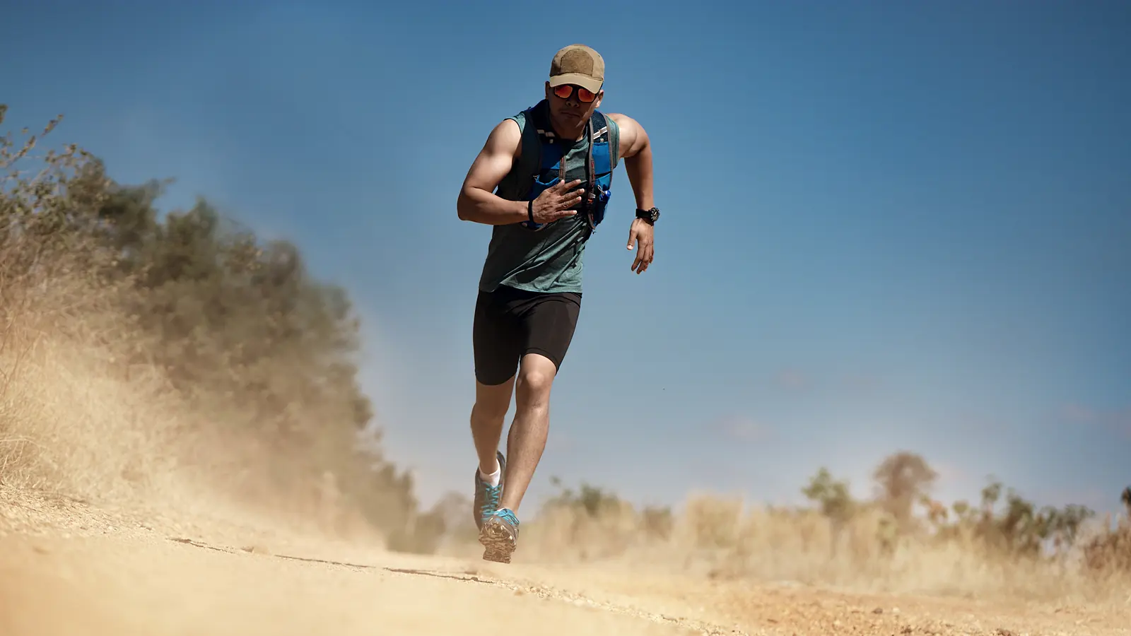 A runner in a singlet on a dry track, kicking up dust in hard sunlight.