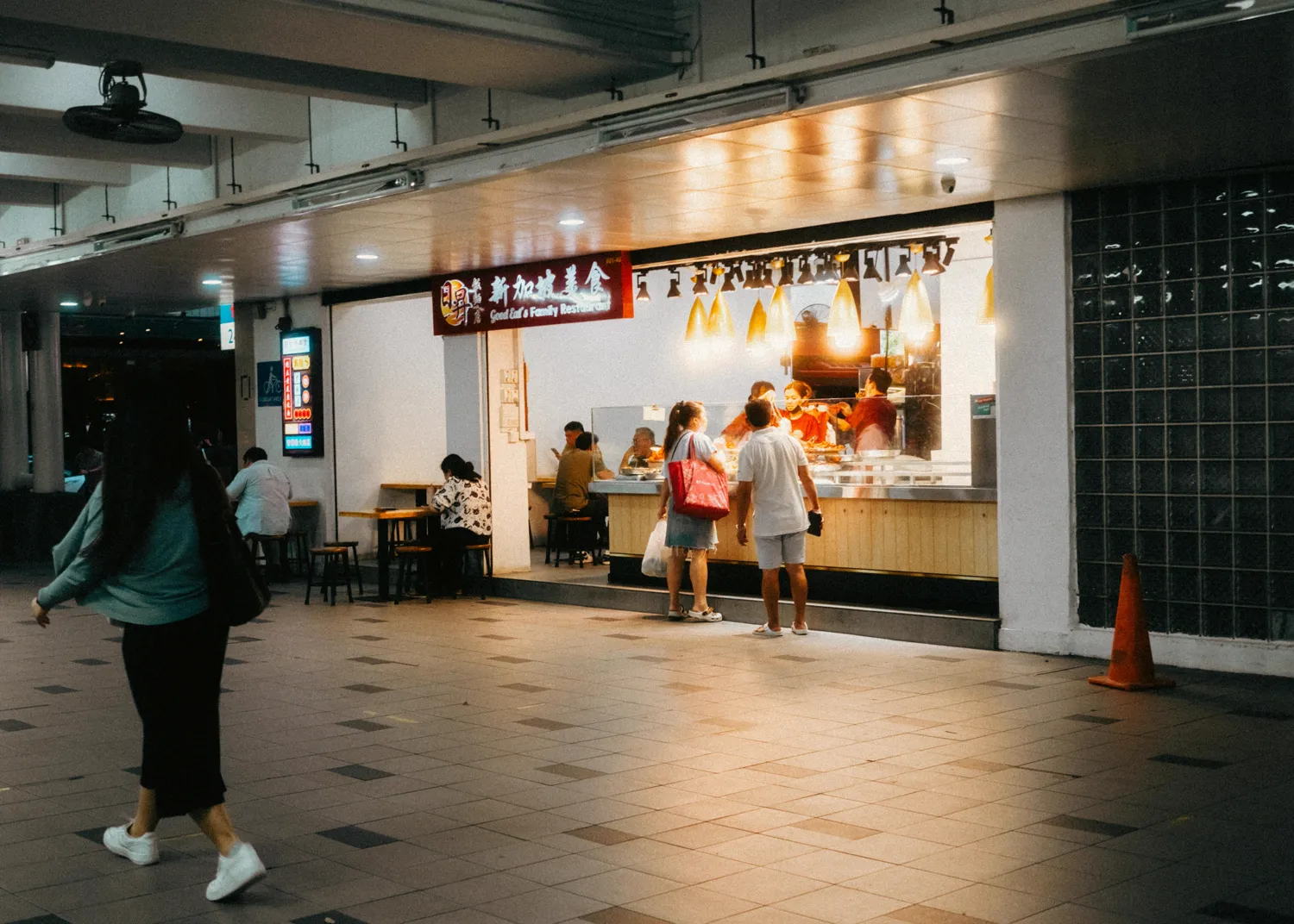 A lit food-court counter at night in Singapore — staff behind the glass, a customer waiting, the walkway dark around it.