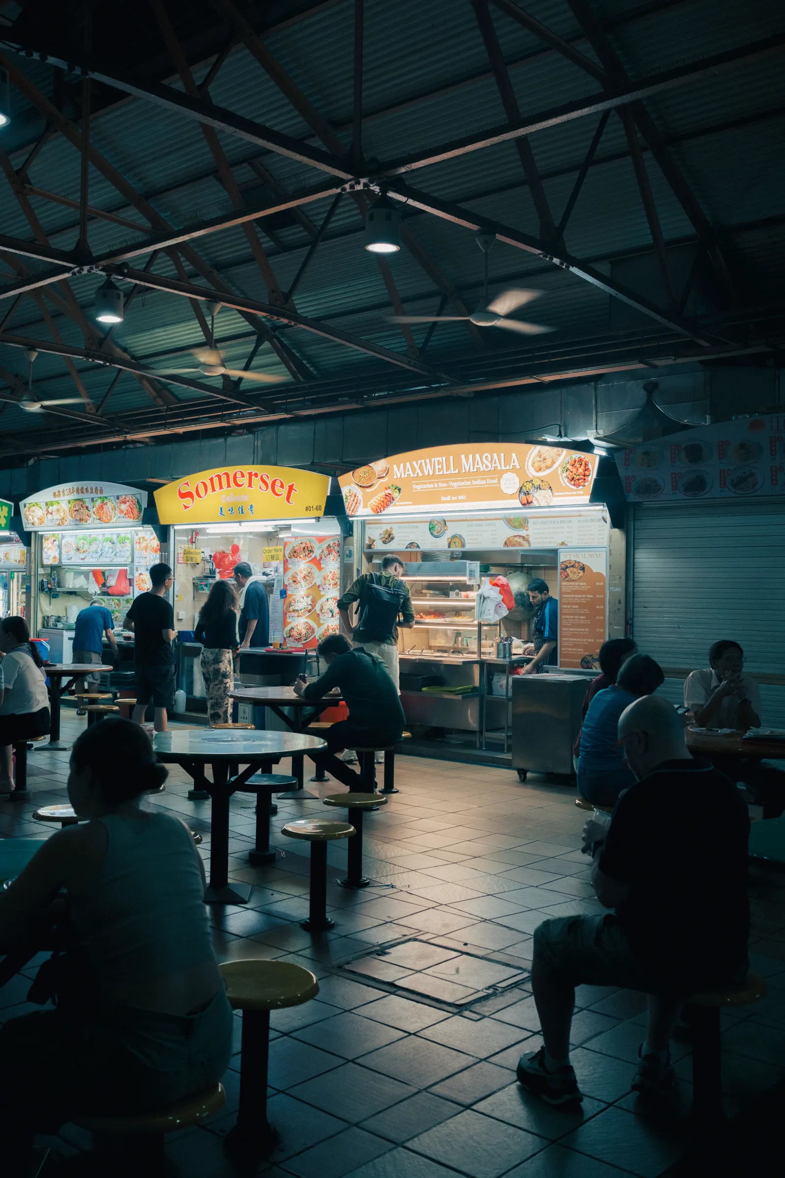 Inside Maxwell Food Centre at night — teal roof trusses and ceiling fans over brightly lit hawker stalls, half the tables still full.