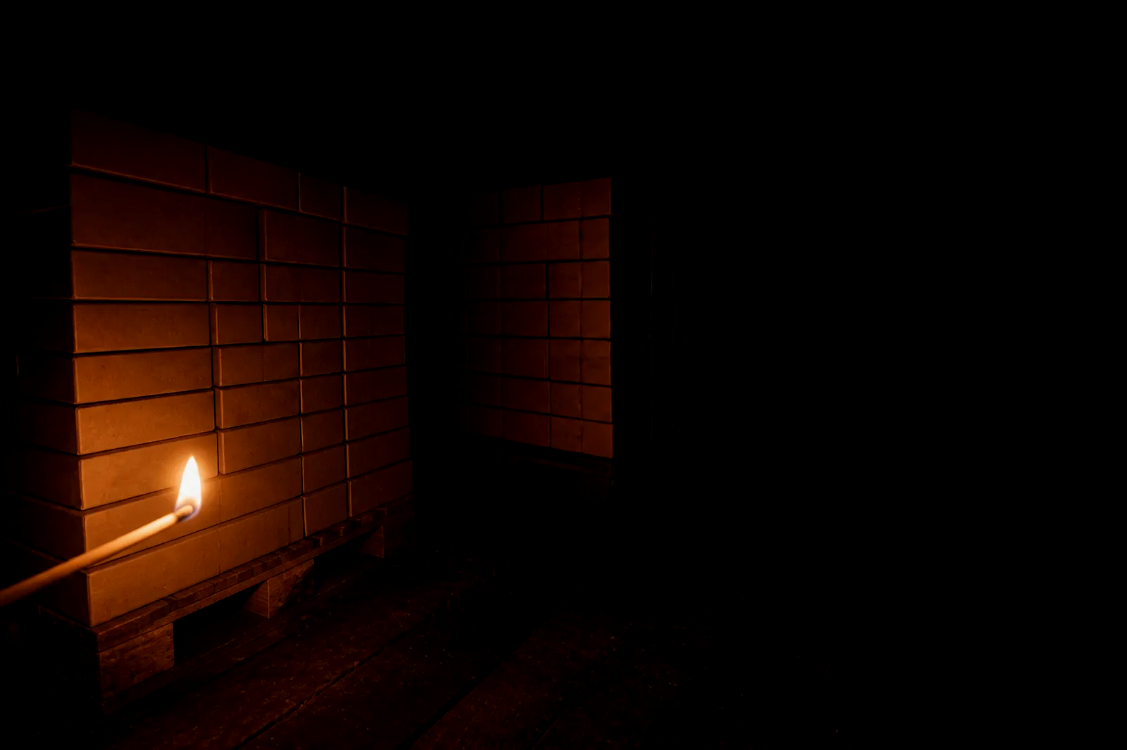 A dark packing aisle of unlabelled cream matchboxes on timber pallets, lit by a single match.